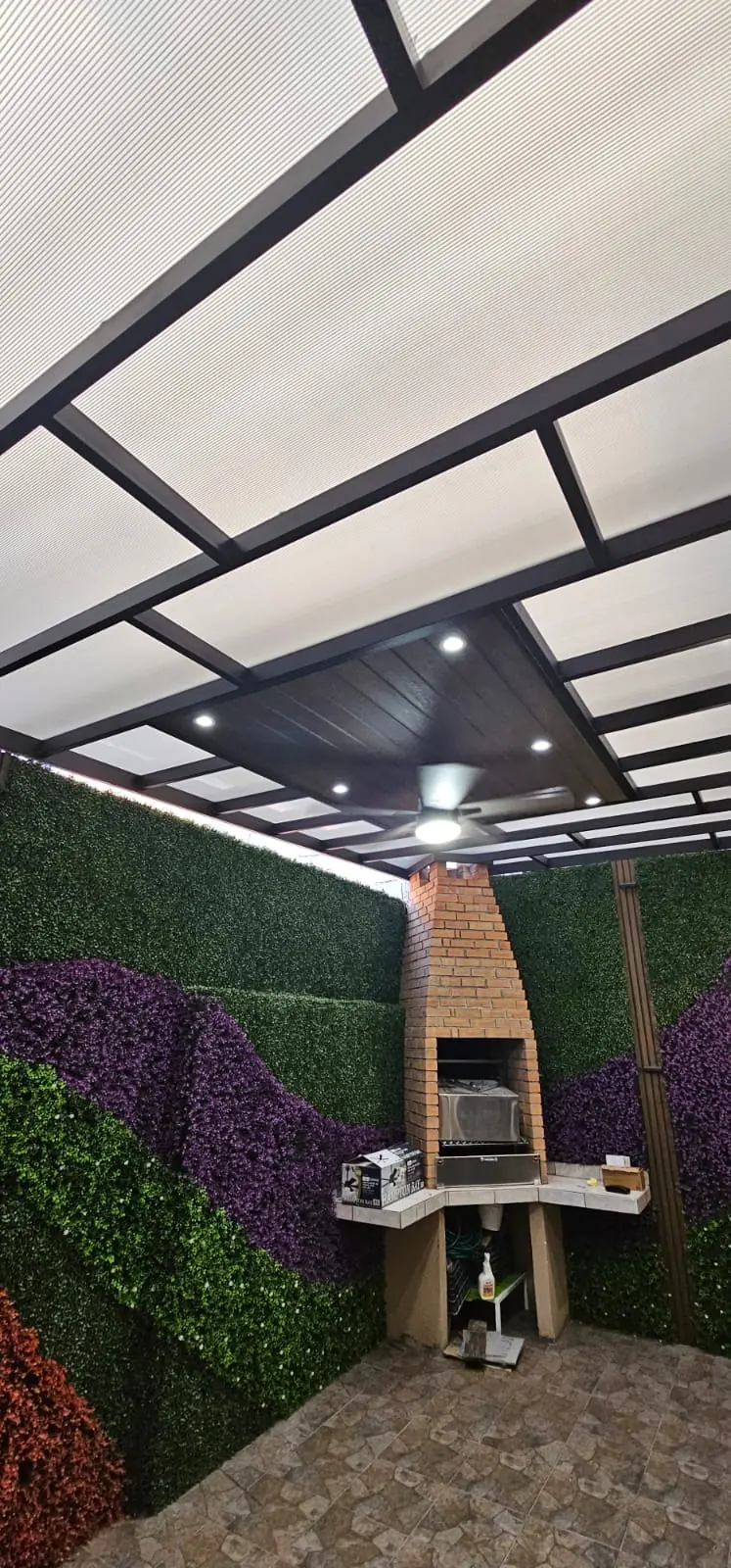 Área de asador cubierta con policarbonato y deck sintético oscuro, junto a un muro verde